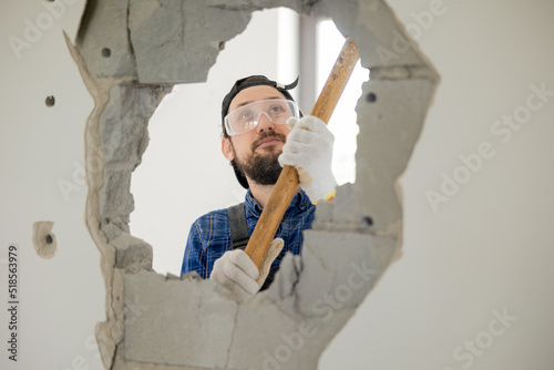 Foto A smiling guy demolishes a wall in a house using a construction hammer on a long stick