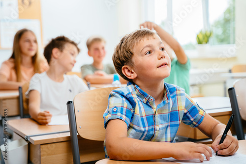 Cute happy blond school boy sitting and listens carefully to the teacher in primary class. Smiling n child pupil studying and writing at school.