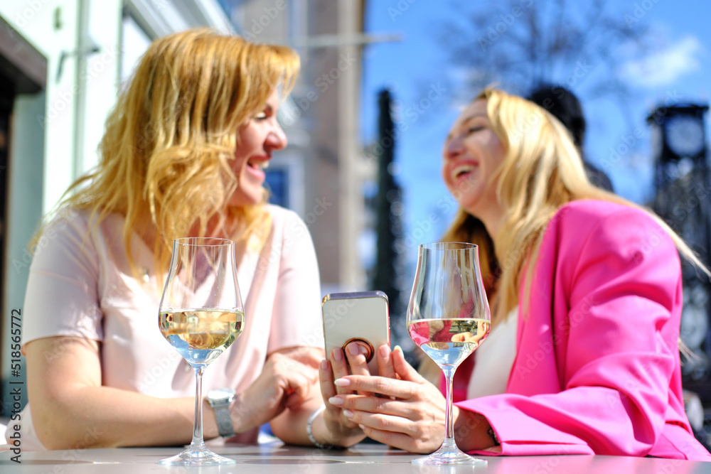 Two adult female friends are sitting at a cafe table on the street in summer, watching funny videos on the phone and laughing Two adult female friends are sitting at a cafe table on the street in summer, watching funny videos on the phone and laughing