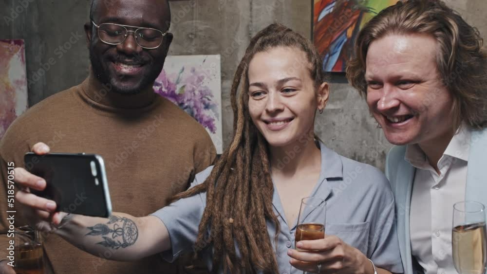 Medium close-up of cheerful Caucasian girl with dreadlocks, businessman and Black man holding glasses of champagne, standing in modern art gallery, taking selfie with smartphone Medium close-up of cheerful Caucasian girl with dreadlocks, businessman and Black man holding glasses of champagne, standing in modern art gallery, taking selfie with smartphone