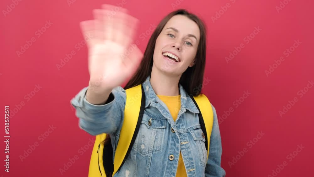 Friendly young student woman saluting with hand, happy expression, wearing denim jacket and yellow backpack, posing isolated over red color background in studio. Education, technology, online concept Friendly young student woman saluting with hand, happy expression, wearing denim jacket and yellow backpack, posing isolated over red color background in studio. Education, technology, online concept