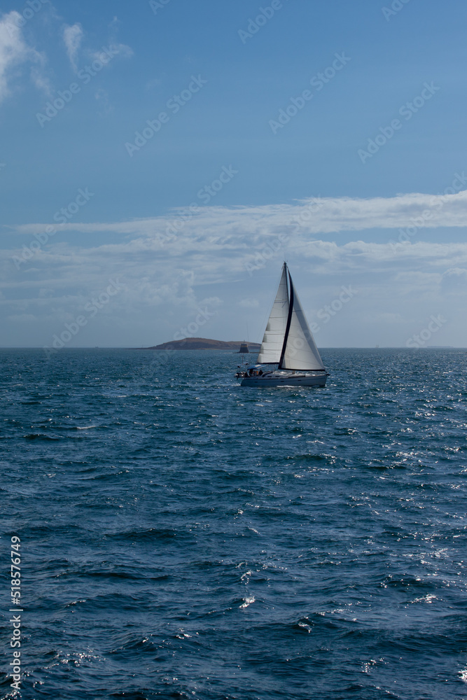 Maritime landscape with a sailboat and an island. A sailboat with a white sail sails in a small sea, behind we can see an island and a signaling buoy. It is the open sea.