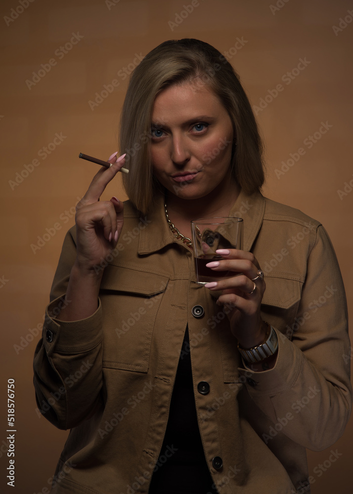 Obraz premium Portrait of a young, beautiful girl with blue eyes and blonde hair in a beige jacket. She looks sternly, smokes a cigarette and holds a glass of whiskey. Expensive watches, gold chain and rings.