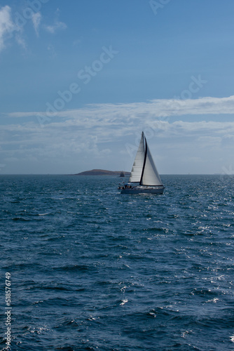 Maritime landscape with a sailboat and an island. A sailboat with a white sail sails in a small sea, behind we can see an island and a signaling buoy. It is the open sea.