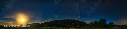 A natural landscape at night. The Milky Way galaxy, the moon, Venus and many stars in the sky above the forest and field