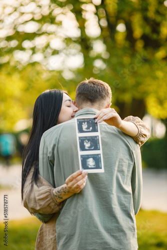 Pregnant woman and her husband standing in a park on a grass and hugging