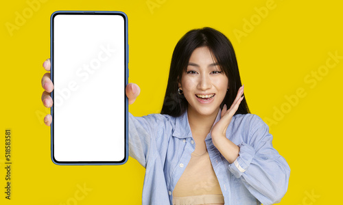 Beautiful asian girl wowing facial expression holding smartphone with white screen happy to introduce new app, game, win, isolated on yellow background. Product placement. Mobile app advertisement