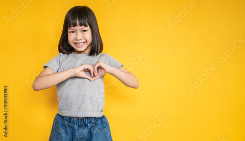 Portrait of young fun smart happy little asian girl isolated on yellow studio shot. Education for elementary kindergarten, little girl hands post in heart shape get back to school, valentines' day