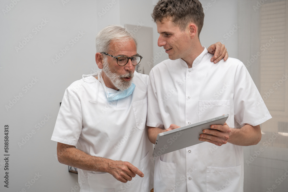 Mature smiling doctor and young intern doctor discussing patient diagnosis, holding digital tablet. Father and son dentist doctors Mature smiling doctor and young intern doctor discussing patient diagnosis, holding digital tablet. Father and son dentist doctors
