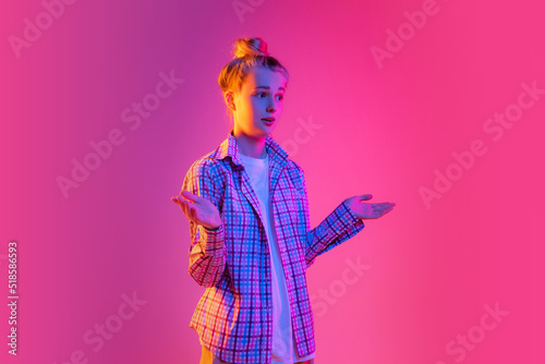 Portrait of adorable young girl, student wearing plaid shirt isolated on magenta color background in neon light. Concept of beauty, art, fashion, emotions