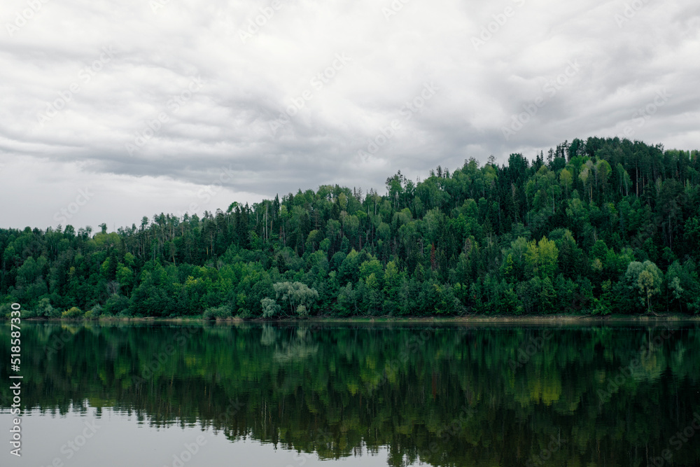Obraz premium Green mixed forest by the lake in the reflection of the water. Calm natural landscape