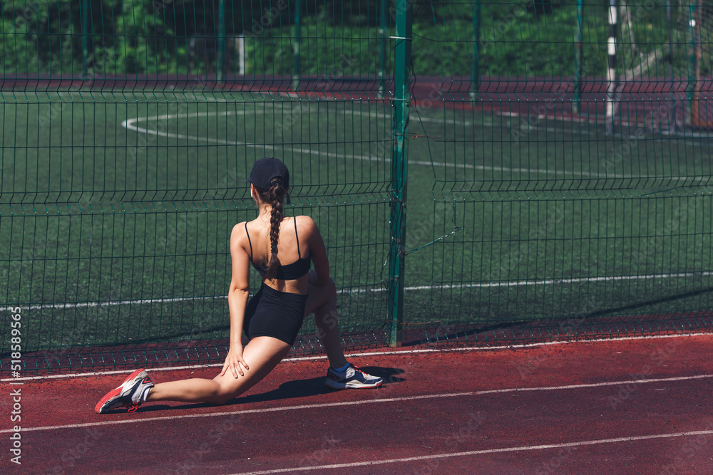young woman doing deep lunge stretching on sports ground outdoors in summer.