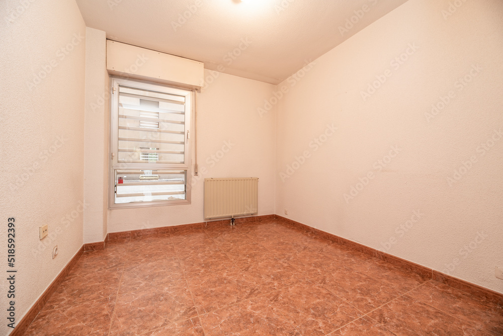 Empty room with reddish stoneware floor, aluminum window with bars and aluminum radiator