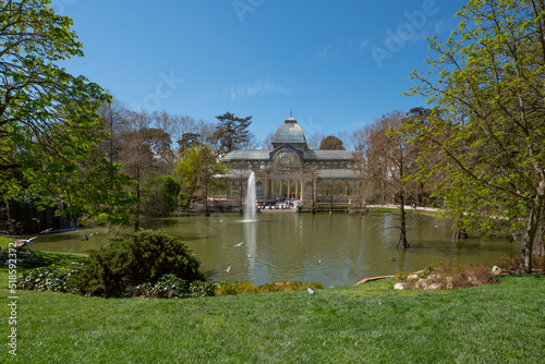 Landscape of the Retiro Park lake with Crystal Palace and trees, lots of green grass in the foreground, water jets and blue skies