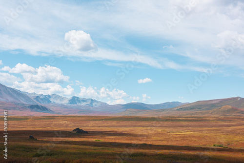 Scenic motley autumn landscape with sunlit high mountain plateau and mountain range under dramatic cloudy sky. Vivid autumn colors in mountains. Sunlight and shadows of clouds in changeable weather.