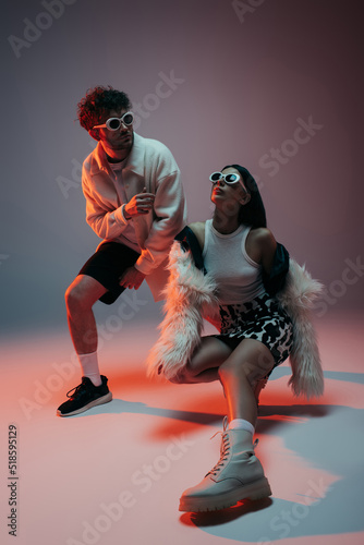 Fototapeta Naklejka Na Ścianę i Meble -  full length of stylish woman in white faux fur jacket and skirt with animal print posing with man in sunglasses on grey.