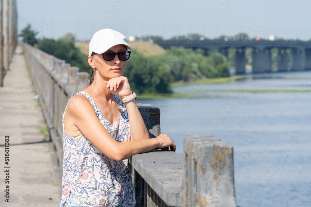 Woman in white cap and sunglasses thoughtfully looks at picturesque river. She stands on the bridge leaning on the iron-concrete handrail. Concept: wonderful moments and instant.