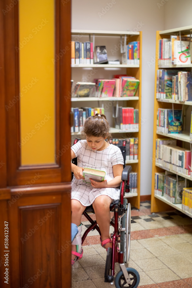 Little girl sits In a wheelchair and reads a book in the library. The life and education of children with special needs. Happy disabled child education concept Little girl sits In a wheelchair and reads a book in the library. The life and education of children with special needs. Happy disabled child education concept