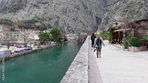 Young curvy woman tourist with backpack walks on sidewalk past river against giant mountains IN old town Kotor, Montenegro