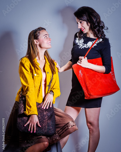 Two young beautiful women, one wearing long sleeve yellow corduroy jacket, dark brown mesh skirt, carrying leather bag, other wearing black dress, shoulder carrying red leather bag, talking other..