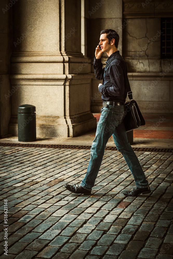 n the City. Young man, shoulder carrying a leather bag, walking on old style street in New York City, talking on cell phone.