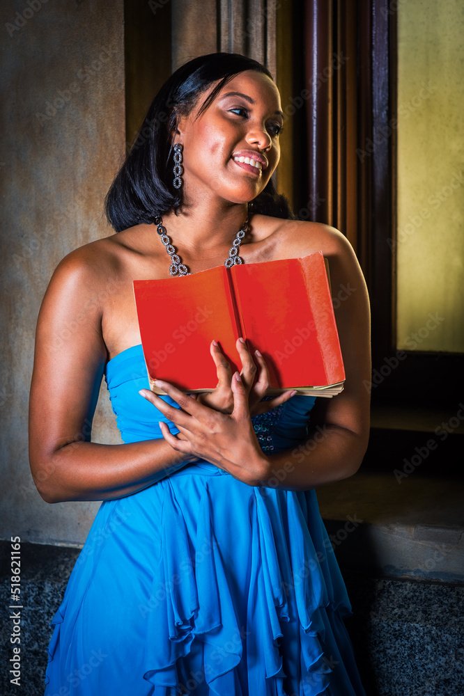 Reading By the Window. Young black woman, holding red book, standing by window, reading, smiling..