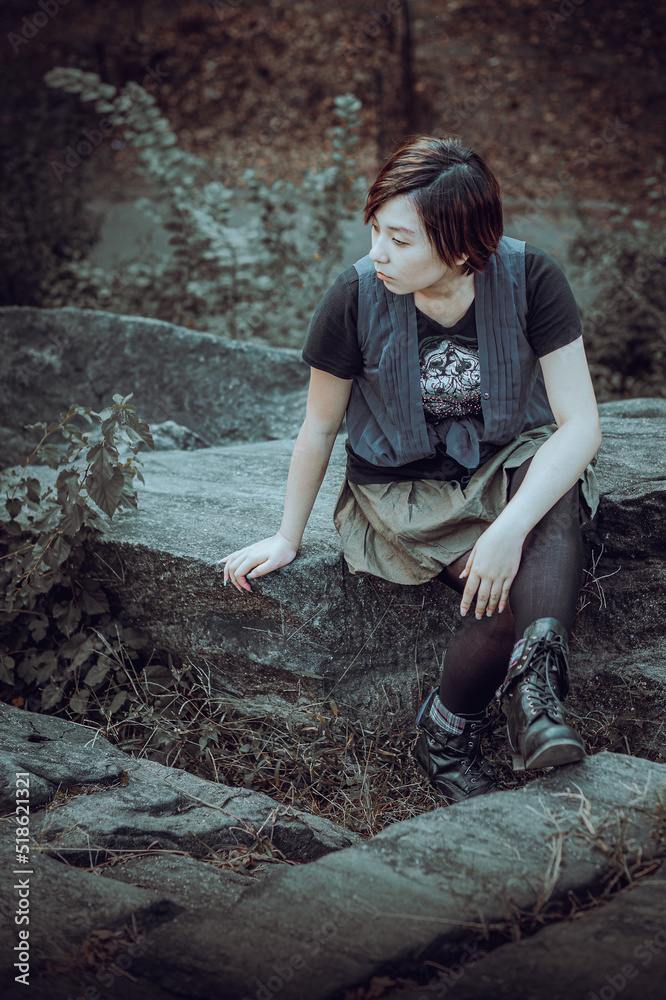 Alone. Young Chinese Girl wearing sleeveless long blouse with black T shirt, black leggings and boots, sitting on rocks, looking around, thinking. Black and White Photography.