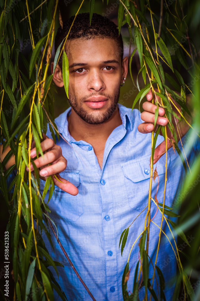 Peek. Young man with little beard, wearing light blue shirt, standing behind long green leaves, thoughtfully looking forward