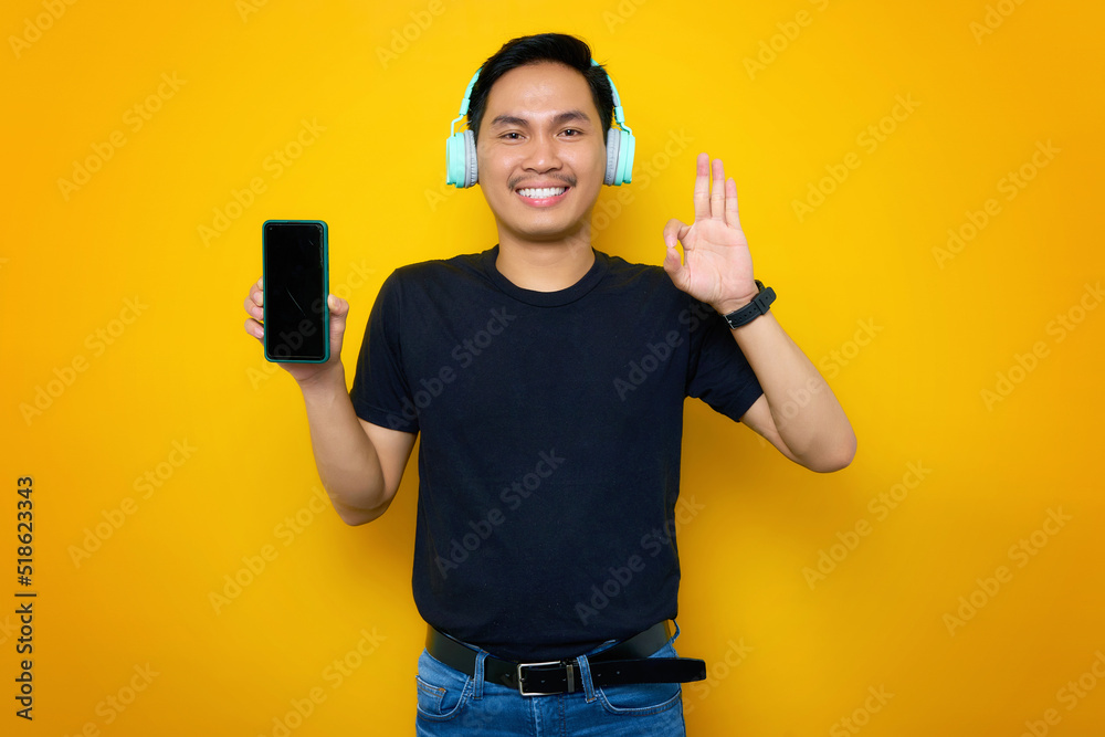 Smiling young Asian man in casual t-shirt with headphones, showing mobile phone with blank screen and showing ok gesture isolated on yellow background Smiling young Asian man in casual t-shirt with headphones, showing mobile phone with blank screen and showing ok gesture isolated on yellow background