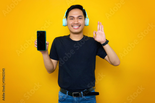 Smiling young Asian man in casual t-shirt with headphones, showing mobile phone with blank screen and showing ok gesture isolated on yellow background