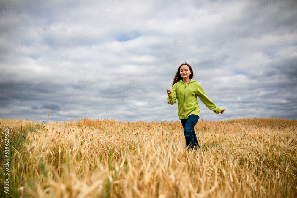 Beautiful smiling preteen girl running against cloudy sky and wheat field in summer