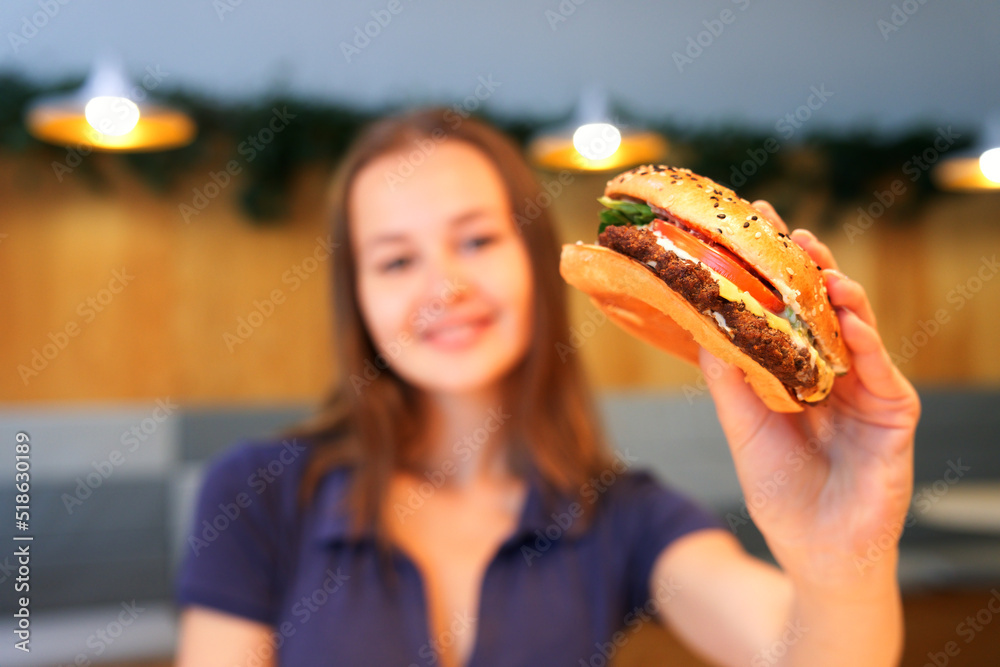 blurred happy positive smiling teen teenager girl, young woman is holding big juicy fat burger, hamburger, Fast food restaurant, junk unhealthy meal concept, selected focus on burger