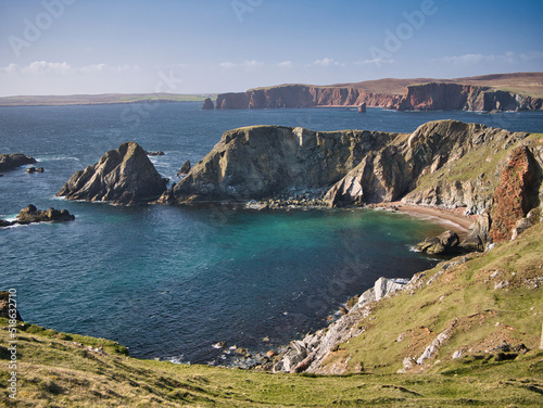 Dramatic coastal cliff scenery and a remote inaccessible beach on the Ness of Hillswick, Northmavine, in the UNESCO Global Geopark of Shetland, UK - taken on a sunny day showing the clear, blue water. photo
