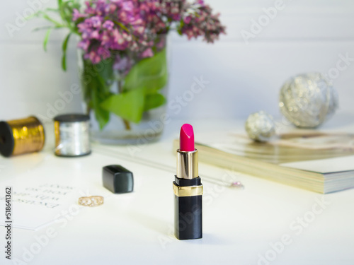 Beautiful female cosmetics like a scarlet red lipstick on the pastel background with silver jewerly, vase of lilac flowers and threds on the table photo