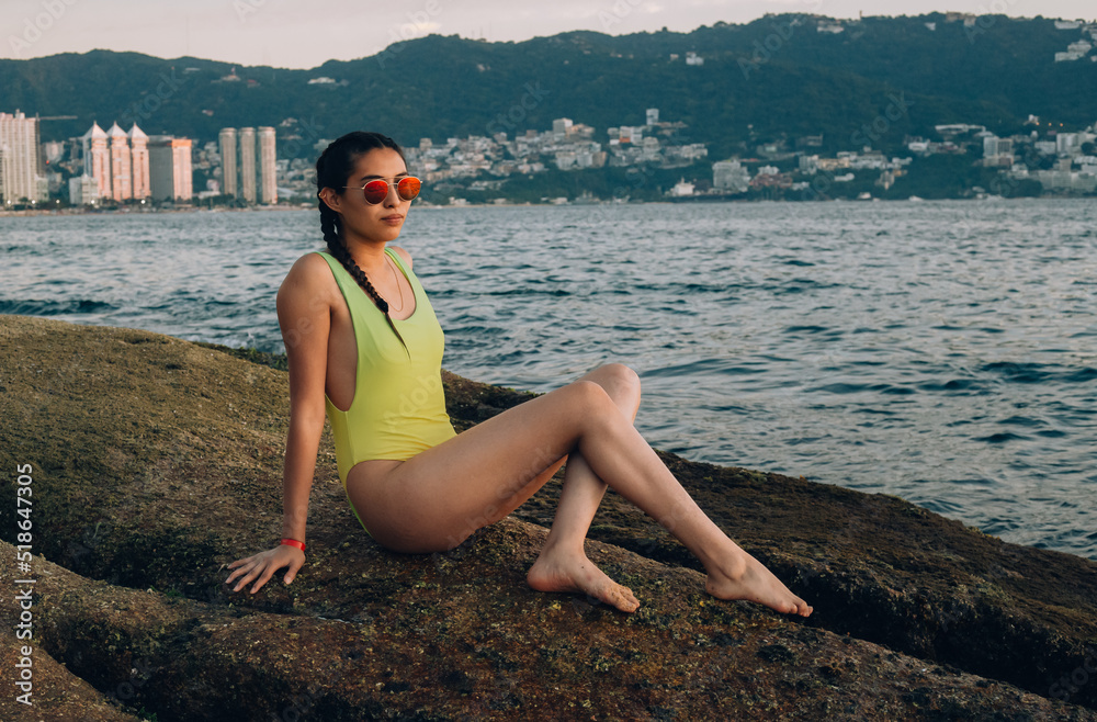 Young pretty generation z girl, with asian mexican appearance and brown skin posing sitting on a rock while enjoying the sea of ​​acapulco at sunset with a gradient yellow swimsuit with orange 