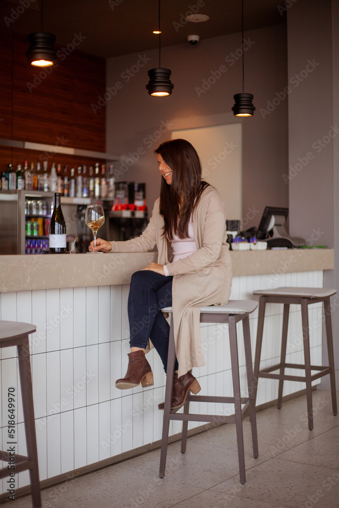 joyful young woman sitting at the bar counter with a glass of wine and a bottle of wine