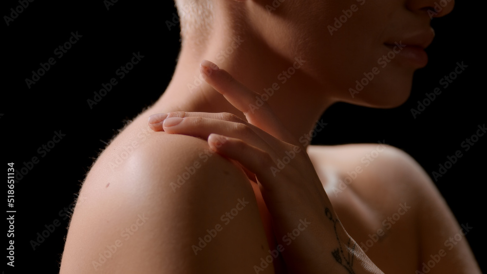 Naklejka premium Big close-up shot of half-turned good-looking young slim white-skinned woman strokes her bare shoulder on black background | Flawless body concept