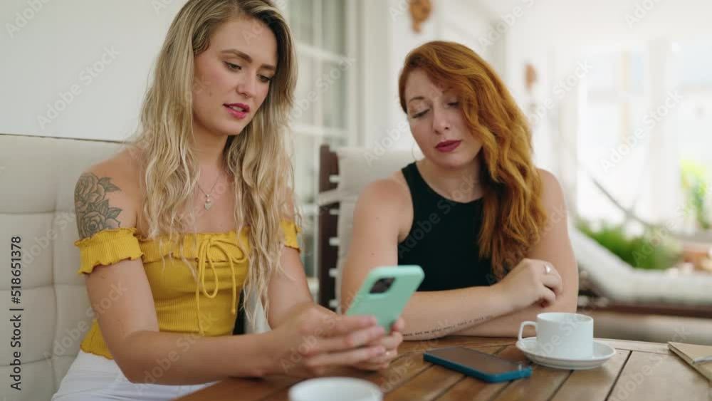 Two women using smartphone and drinking coffee sitting on table at home terrace Two women using smartphone and drinking coffee sitting on table at home terrace