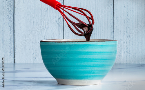 Teal blue bowl on a light table and background with a red whisk dripping melted chocolate 