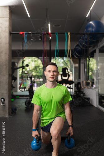 A young man exercises and performs knee and leg exercises, split, squat, holding the kettlebells with his hands, at the local training and fitness center