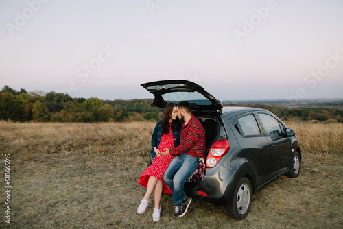 Couple of newlyweds sit in the trunk of a new family car. A pregnant woman in a red dress and denim jacket cuddles with her husband. Autumn landscape. High quality photo