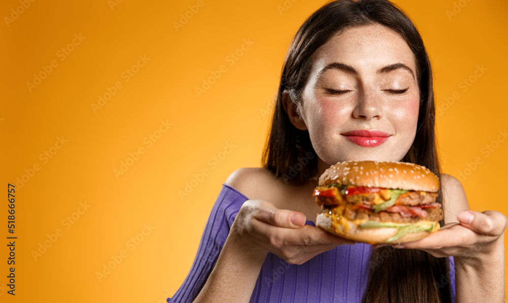 Smiling woman smells her tasty fresh burger from restaurant delivery. Girl holds cheeseburger on plate and sniff it, stands over orange background
