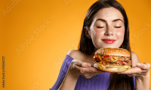 Smiling woman smells her tasty fresh burger from restaurant delivery. Girl holds cheeseburger on plate and sniff it  stands over orange background