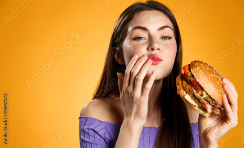 Chewing woman holding tasty fast food burger  cleaning her mouth after taking a bite. Satisfied girl eats her delivery meal  orange background