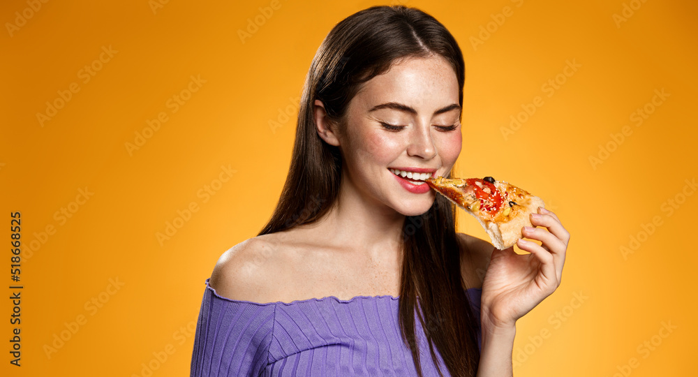 Image of girl smiling while biting slice of pizza, happy face at pizzeria restaurant, orange background Image of girl smiling while biting slice of pizza, happy face at pizzeria restaurant, orange background
