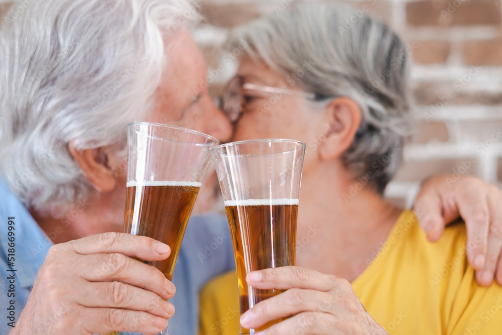 Defocused senior couple kissing holding two glasses of beer, brick wall background Defocused senior couple kissing holding two glasses of beer, brick wall background