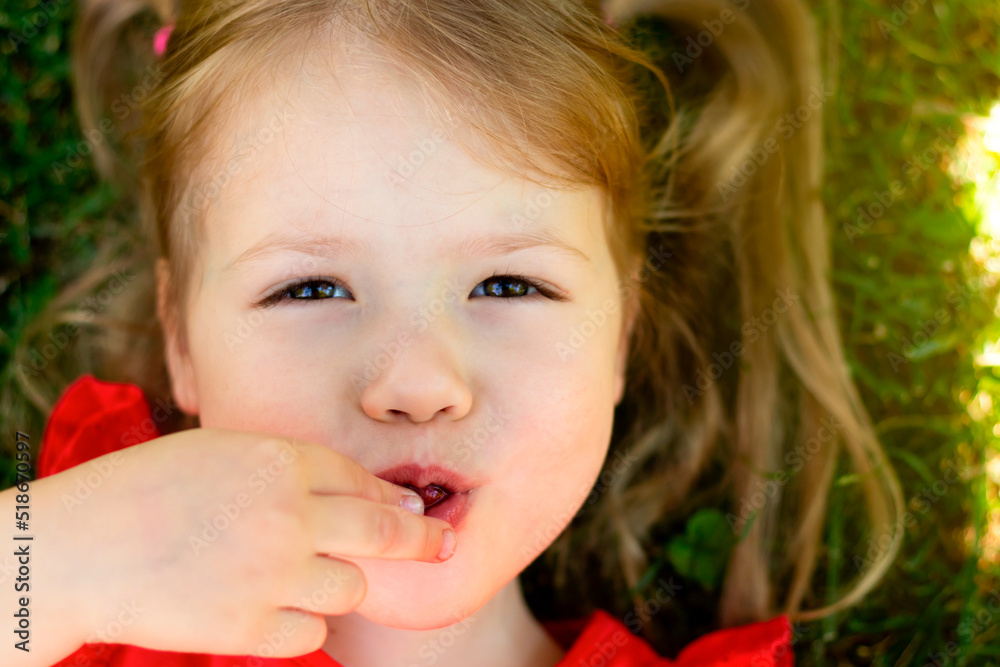 Close-up of face of smiling caucasian girl eating cherries lies on the grass in the garden in summer Close-up of face of smiling caucasian girl eating cherries lies on the grass in the garden in summer