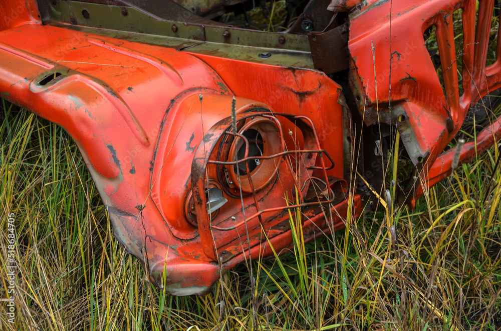 Headlight and front metal grill of old rusty red truck at junkyard vehicles in overgrown weedy area Headlight and front metal grill of old rusty red truck at junkyard vehicles in overgrown weedy area