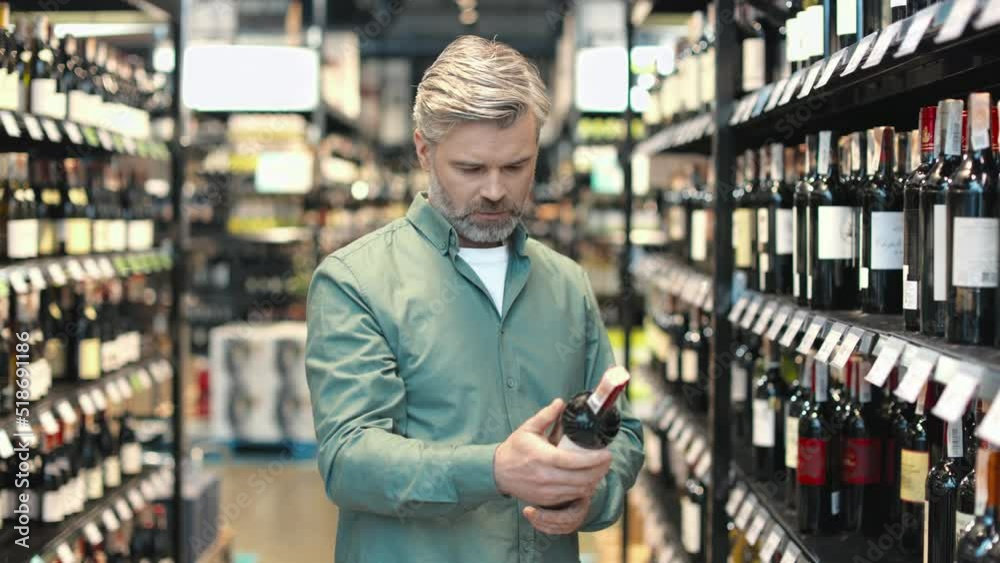 Mature man choosing red wine while standing in front of shelves full of bottles in alcohol market. Handsome male buyer reading label and deciding which kind to buy. Mature man choosing red wine while standing in front of shelves full of bottles in alcohol market. Handsome male buyer reading label and deciding which kind to buy.