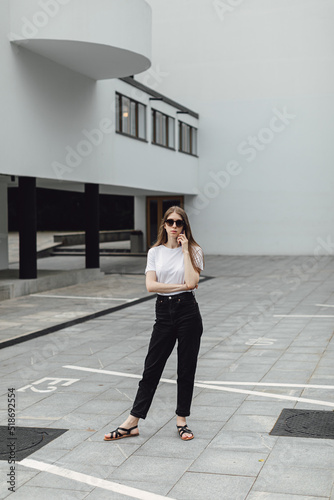 Young woman in white t-shirt standing in front of grey building and using smartphone. Urban style and street fashion. Girl in sunglasses with phone. Chatting, messaging, reading